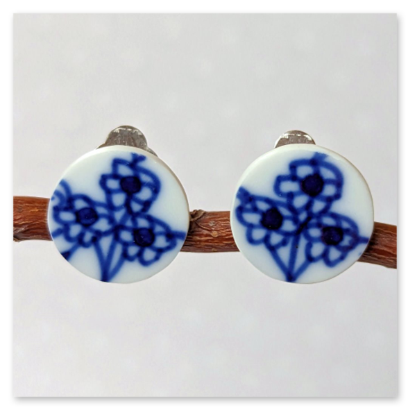 Runde Ohrclips aus den Scherben eines alten Tellers hergestellt. Das Motiv zeigt jeweils drei blaue Blüten an einem gemeinsamen Stiel. Die kräftige blaue Farbe erinnert an traditionelle Porzellanmalerei.