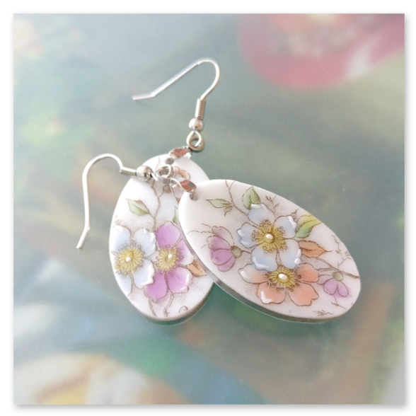 Hängeohrringe mit silberfarbenen Haken aus Edelstahl. Die Anhänger sind oval und bestehen aus weißem Porzellan mit zarten Blumenmotiven in Pastellfarben.