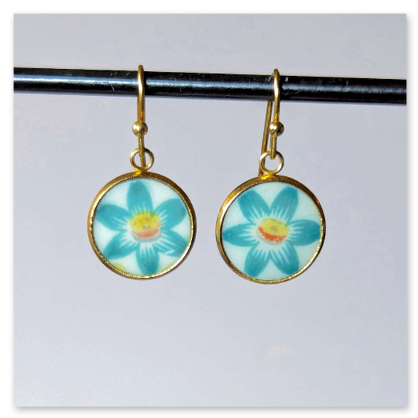 Diese kleinen filigran gearbeiteten Ohrhänger bestechen durch ihr frisches, florales Design. In runden goldfarbenen Fassungen sind jeweils ein zartes Blütenmotiv eingefasst, deren türkisfarbene Blätter sich harmonisch um ein warmes, gelb-orangefarbenes Zentrum gruppieren. Die leicht glänzende Porzellanoberfläche  lässt die Farben lebendig wirken.