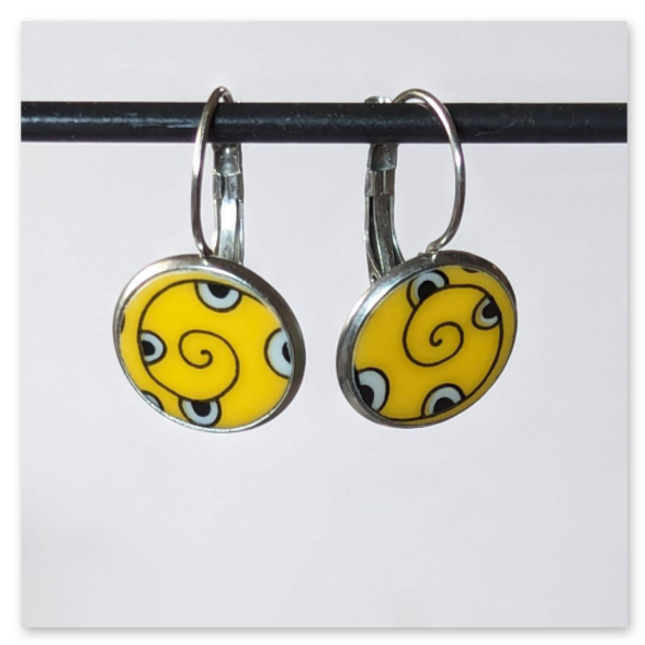 Diese besonderen Ohrhänger sind echte Unikate. Gefertigt aus einer Scherbe alten Porzellangeschirrs, vereinen sie Nachhaltigkeit mit individuellem Design. Das auffällige gelbe Muster mit spiralförmigem Motiv macht jedes Paar zu einem kleinen Blickfang.<br /><br />Die Fassungen sowie die praktische Klappbrisur bestehen aus robustem Edelstahl und sind angenehm zu tragen.