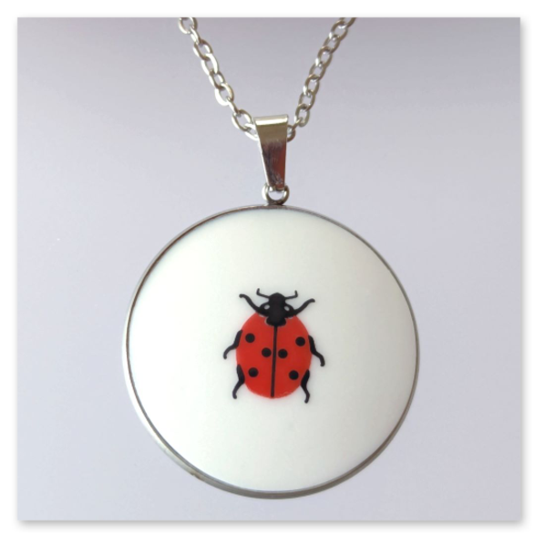 Ein runder Edelstahl-Kettenanhänger mit einem Durchmesser von 30 mm, in dem ein Porzellanstück mit Marienkäfermotiv zu sehen ist.  Das Porzellan einer Tellerscherbe wurde geschliffen und passgenau in die silberfarbene Fassung eingesetzt.