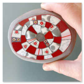 Das handgefertigte Mosaik auf dem Naturstein vereint Porzellan, Farbglas und eine kleine Metallspirale zu einem kunstvollen  Motiv.<br />Der Name "Harmaan Punainen" bedeutet übersetzt "Grau-Rot" und beschreibt somit perfekt die Farbkombinationen des Mosaiks.